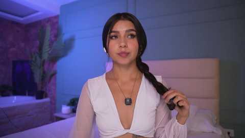 Video of a young woman with long dark hair in a braid, wearing a white crop top and headphones, standing in a modern bedroom with blue and purple lighting, and a white headboard.