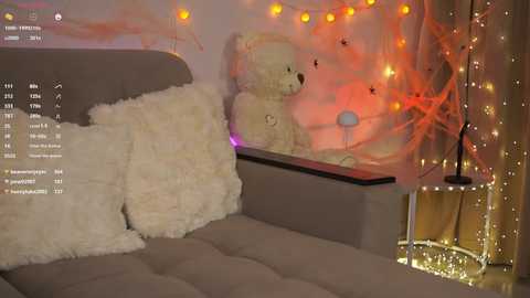 Video of a cozy room with a gray couch, a fluffy white pillow, a large teddy bear, and warm, glowing fairy lights.