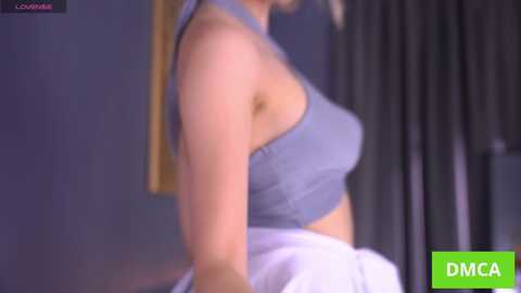 A video of a fair-skinned woman with blonde hair, wearing a light blue sleeveless top, and white skirt, sitting on a bed. Background features dark curtains and a framed picture.