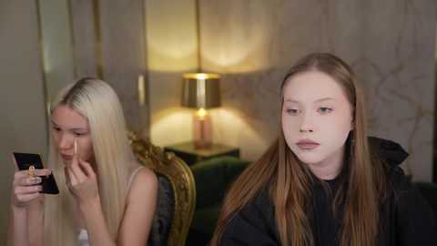 Media: Video of two young women seated on a green velvet couch in a dimly lit room. One, with long blonde hair, focuses on a smartphone. The other, with long brown hair, wears a black hoodie and stares pensively.