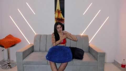 Video of a dark-haired woman with fair skin, wearing a red bra and blue skirt, seated on a gray sofa with a black pillow, surrounded by modern, white, angular light fixtures.