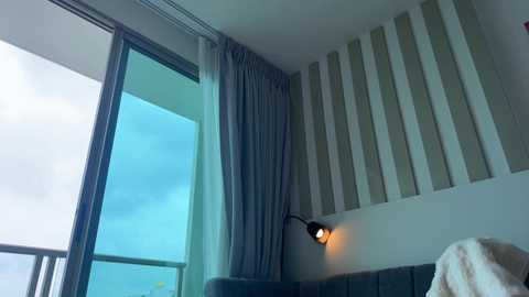 Video of a modern hotel room with a large glass window overlooking a cloudy sky, featuring a wall-mounted sconce and a beige curtain. The room is minimalist, with a gray upholstered headboard and a textured white wall.