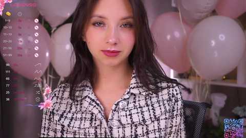 Video of a young woman with long dark hair, fair skin, and red lipstick, wearing a black-and-white checkered blouse. Background features pink balloons and a calendar.