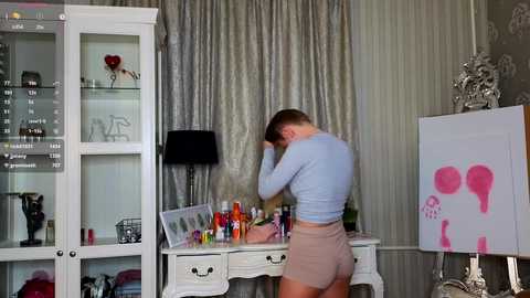 Media: Video of a man in a light blue shirt and beige shorts painting a woman's face on a white table, surrounded by makeup and pink heart-shaped brushes.