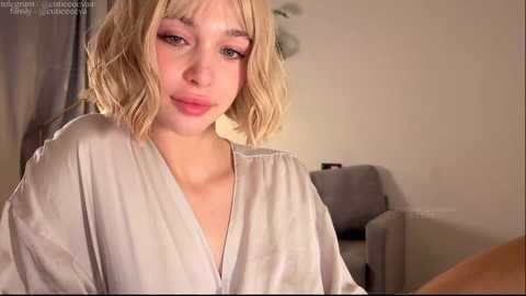 A video of a young woman with fair skin and shoulder-length, wavy blonde hair, wearing a light-colored blouse. She has a slight smile and is sitting indoors, with a grey sofa and beige wall in the background.