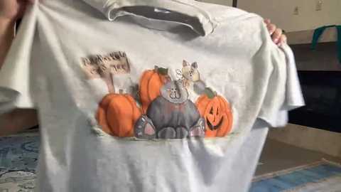 Video of a person holding a white t-shirt featuring a cartoon cat in a chef's hat, surrounded by pumpkins. Background includes a kitchen with dark countertops and beige walls.