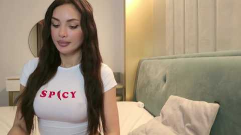 Video of a young woman with long black hair, fair skin, wearing a tight white t-shirt with \"SPICY\" printed in red, in a modern bedroom with a light blue upholstered headboard.