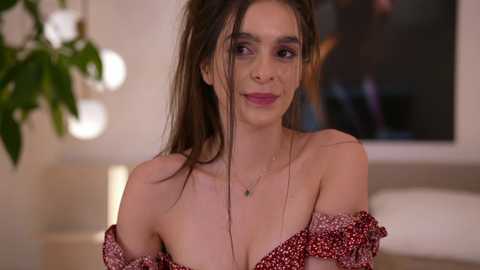 Video of a young woman with fair skin, long brown hair, and a slight smile, wearing a red off-shoulder dress, standing in a softly lit room with a leafy plant and a large window in the background.