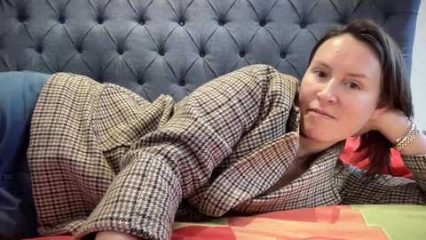 Media: A video of a woman lying on a bed with a blue, tufted headboard, wearing a plaid coat, a red shirt, and a bracelet. She has a neutral expression.