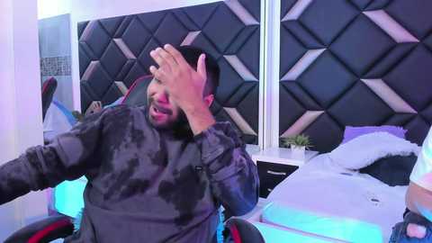 Video of a man with a beard, wearing a camo hoodie, sitting in a chair, covering his face with his hand, in a modern bedroom with a diamond-patterned headboard.