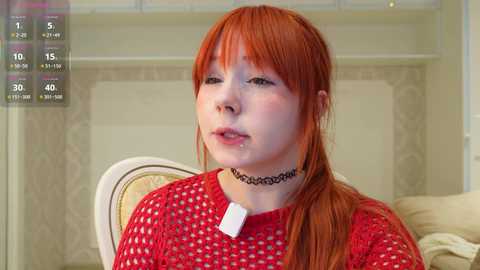Video of a young, pale-skinned woman with red hair in a ponytail, wearing a black choker and red knit sweater, seated indoors with a beige cushion behind her.