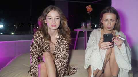 Video of two women with fair skin and light brown hair, one wearing a leopard print robe, the other a cream cardigan, seated indoors at night, holding a phone.