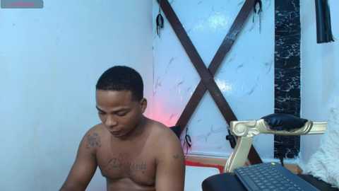 Video of a shirtless Black man with tattoos, sitting on a chair in a dimly lit room with a marbled wall, a black keyboard, and a gold-framed mirror.