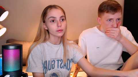 Video of a blonde woman wearing a \"Paul T. Shirt\" t-shirt, sitting next to a boy with short hair, both looking serious in a dimly lit room with a lamp and speaker.