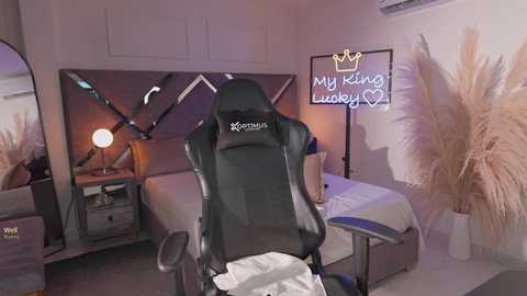Video of a cozy, dimly-lit gaming room featuring a black gaming chair, a wooden bed with white linens, a desk lamp, a neon sign reading \"My King Lucky \u2665,\" and beige decorative plants.