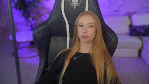 Video of a young, slender, Caucasian woman with long, straight blonde hair, wearing a black jacket, seated in a black gaming chair with white accents in a dimly lit, modern living room with purple lighting.