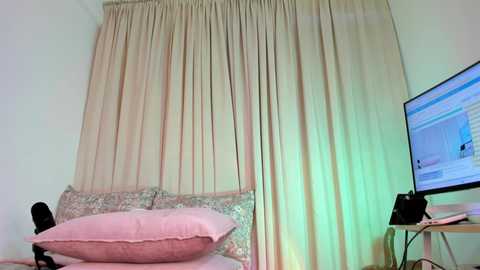 Video of a bedroom with beige curtains, pink pillows, a floral-patterned bedspread, and a black computer monitor displaying a website on a wooden table.