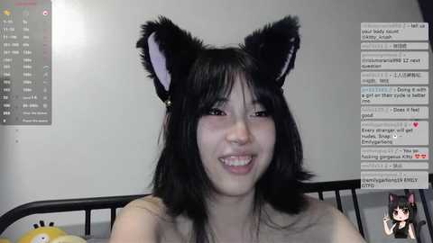 Video of a smiling Asian woman with long black hair wearing cat ears, seated on a bed. Background includes a gray wall and a chat window displaying messages.