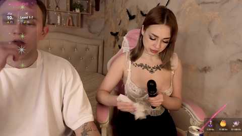 Media: Video of a light-skinned woman with long brown hair, wearing a revealing lace top and black skirt, using a vibrator on a man in a beige chair in a dimly lit, feminine room.