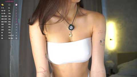 Media: Video of a young woman with straight, dark brown hair, wearing a white, strapless bandeau top, multiple tattoos, and a gold necklace with a black disc pendant, standing in a dimly lit room with a soft light source.