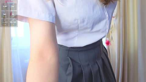A video of a woman wearing a white blouse and pleated grey skirt, with a partially visible arm and a pink flower in her hair. Background shows light-colored curtains.
