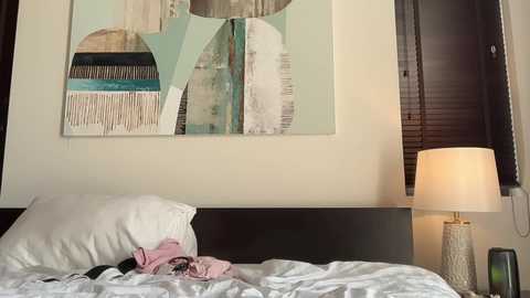 Video of a modern bedroom with a dark wooden headboard, white bed linens, a pastel painting, and a beige lamp on a nightstand.