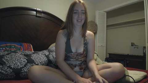 Video of a fair-skinned, slim woman with long red hair, wearing a floral crop top, meditating cross-legged on a bed with colorful bedding in a dimly lit bedroom.