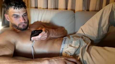 Video of a shirtless, muscular, light-skinned man with short dark hair and a beard, wearing beige Calvin Klein underwear, lying on a beige couch, engrossed in a smartphone.