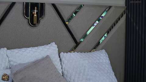 Video of a modern bed with a geometric-patterned headboard, beige upholstery, and a light gray pillow. A small, decorative oil lamp hangs on the wall above.