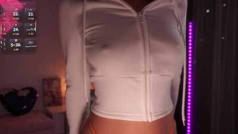 Video of a woman in a white zip-up hoodie, highlighting her medium-sized breasts through the fabric. Background shows a dimly lit room with a TV screen displaying a video game.