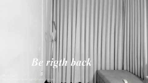 Black-and-white video of a minimalist bedroom with a single bed against a wall of vertical, pleated curtains. A lamp stands beside the bed, and the text \"Be right back\" is overlaid on the image.