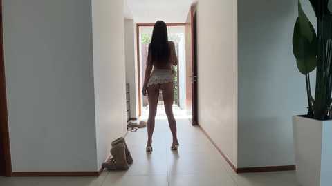 Media: Video of a slender woman with long hair, wearing a white lace bra and panties, standing in a minimalist hallway with white walls, wooden doors, and a potted plant.