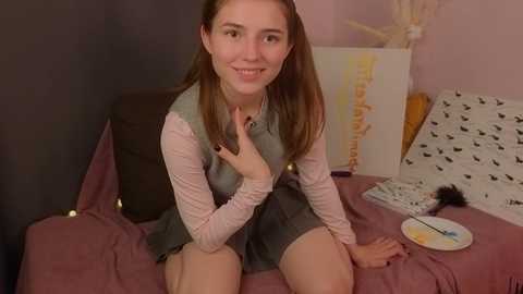 Media: Video of a young woman with light skin, straight brown hair, and a slight smile, sitting on a bed with a pink comforter. She wears a grey sleeveless dress over a long-sleeve white shirt. The room has a soft pink wall and various art supplies scattered around.