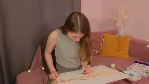Media: Video of a woman with long brown hair, wearing a light blue sleeveless top, painting on a pink cushion in a pink room with gray curtains and yellow pillows.
