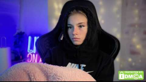 Video of a young woman with light skin, dark hair, and serious expression, wearing a black hooded sweatshirt, sitting on a bed with a pink blanket. Background features a soft, glowing fairy light string and a neon sign partially visible.