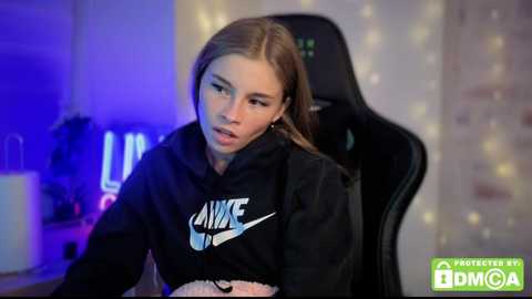 A video of a young woman with light skin and long brown hair, wearing a black Nike hoodie, seated in a gaming chair. The background features a soft, warm glow and gaming equipment.