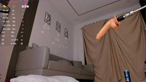 A video of a modern living room with a grey couch, beige curtains, and a penis-shaped object on a pole. The room features black-and-white framed art and a video game interface in the top left corner.