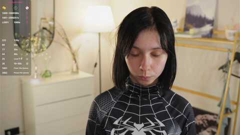 Video of a young Asian woman with straight black hair, wearing a black Spider-Man suit, standing in a softly lit bedroom with a white dresser, lamp, and a framed landscape painting in the background.