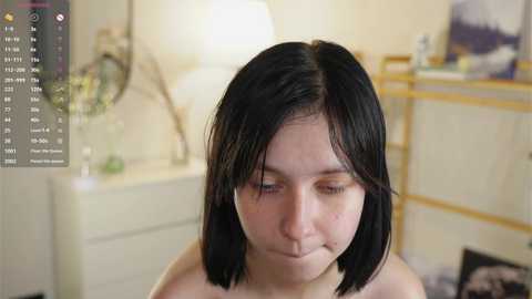 A video of a young, fair-skinned woman with shoulder-length black hair, wearing a black bra, in a dimly lit bedroom.