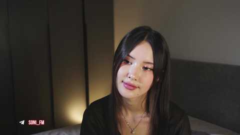 Video of an East Asian woman with long, straight black hair, wearing a black top, sitting on a bed in a dimly lit room.