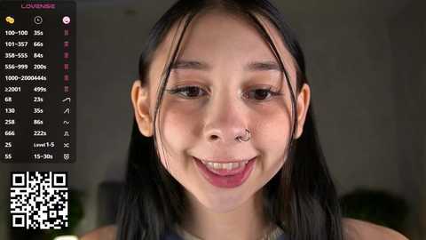 Video of an Asian woman with long black hair, light skin, and a nose piercing, smiling in a room with a blurred background.