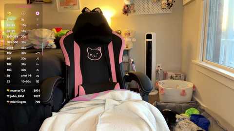 Video of a gaming chair with a black and pink design, featuring a cat head logo, in a cluttered room with laundry baskets, toys, and a window.