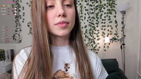 Media: Video of a young woman with long, straight brown hair, fair skin, and a neutral expression, wearing a white t-shirt with a deer graphic. Background includes green ivy wall, white lamp, and green couch.