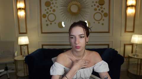 Video of a young woman with pale skin, brown hair in a bun, wearing a white off-shoulder top, sitting on a dark blue couch in a luxurious room with gold and white decor, including lamps and a circular sunburst mirror.