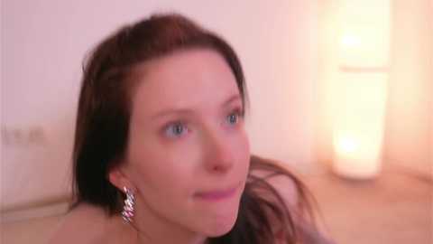 Video of a Caucasian woman with long brown hair, blue eyes, and light skin, wearing silver earrings. She has a calm expression and is indoors with soft, warm lighting.