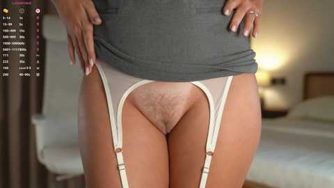 Video of a woman lifting a gray top, revealing white garter belt with visible pubic hair, tan skin, hands on hips, blurry bedroom background.