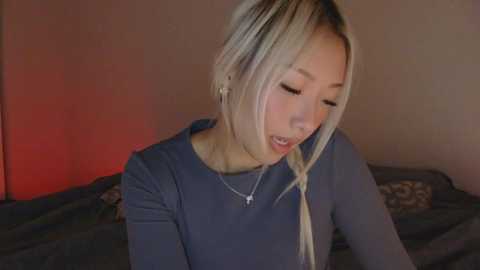 A video of a young Asian woman with platinum blonde hair, wearing a gray long-sleeve top, sitting on a bed with patterned bedding. The background has a red light effect, creating a warm ambiance.