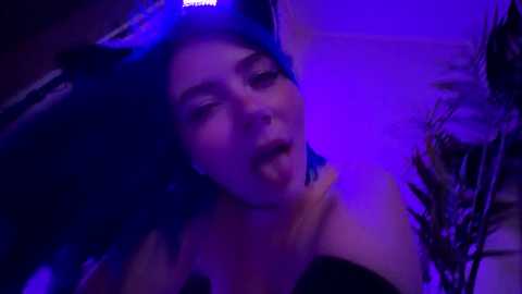 Media: A dimly lit video of a young woman with pale skin, blue hair, and a black headband, wearing a black top, with a purple glow highlighting her face and tongue sticking out.