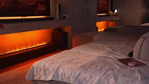 Media: Video of a dimly-lit bedroom with a large, cozy fireplace beneath a window, casting a warm glow on the bed with white linens and a red book.