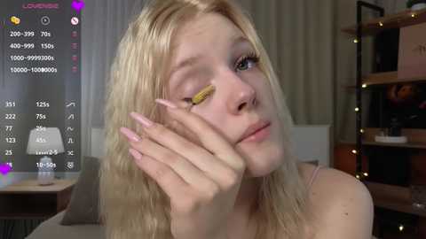 Video of a young blonde woman with long hair, wearing a yellow eye patch and pink manicured nails, in a cozy indoor setting.